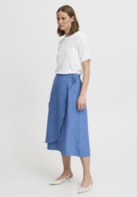 Hvid, tekstureret bluse med korte, flæseærmer og en blå chambray omslagsnederdel. Outfitet er parret med hvide spidse hæle.