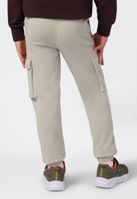 Person som har på seg beige cargo-bukser og olivengrønne joggesko med hvite såler, står med hendene nær sidelommene, vendt bort.