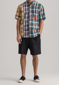 Camisa de manga corta de cuadros multicolores que presenta patrones variados en amarillo, naranja y verde, combinada con pantalones cortos negros y zapatillas.