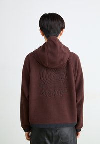 Chaqueta de forro polar marrón con capucha, que presenta un patrón texturizado en la parte posterior y un dobladillo de tela contrastante. Diseño simple y funcional.