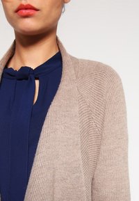 Cardigan beige a maglia con tessuto testurizzato, apertura davanti e bordi leggermente costolati sopra una blusa blu navy con dettaglio colletto a fiocco.