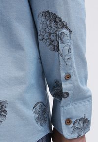 Lichtblauwe blouse met lange mouwen, voorzien van zwart gedetailleerde bloemen- en botanische prints, bruine knopen op de manchetten en mouwsplit.