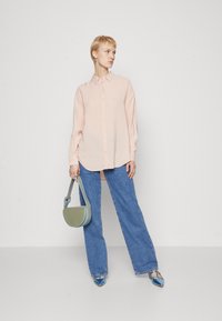 Camicia button-up rosa chiaro con maniche lunghe, abbinata a jeans blu a gamba larga. Gli accessori includono una borsa verde e scarpe con tacco blu.