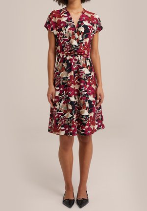 Kvinde iført en knælængde, kortærmet blomsterkjole i rød, beige og sort, og sorte spidse flade sko.