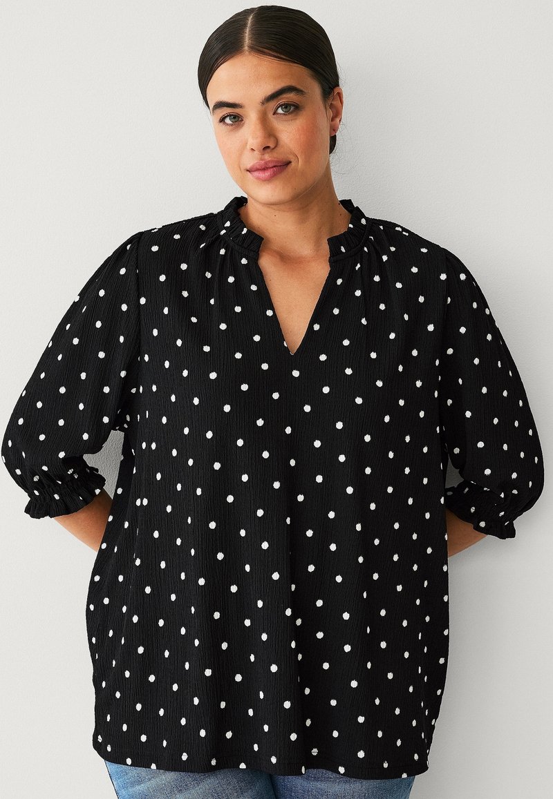 Black polka dot blouse with a V-neck, textured fabric, puffed sleeves, and gathered neckline, featuring white spots evenly distributed.