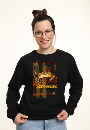 Zwarte sweatshirt met een afbeelding van een krab en de tekst "NORTH PACIFIC" in oranje en geel, tegen een gestructureerde achtergrond in gedempte kleuren.