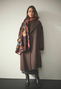 Brun frakke med store revers, parret med et blomsterpræget tørklæde i burgunder, båret over en mørk sweater og grå nederdel, komplet med sorte støvler.