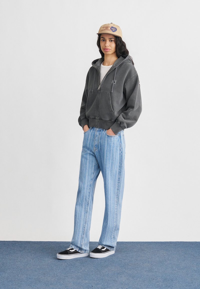 Giovane con un berretto beige, felpa grigia con zip, jeans a righe azzurri chiari e scarpe da ginnastica nero-bianco, in piedi su un tappeto blu.