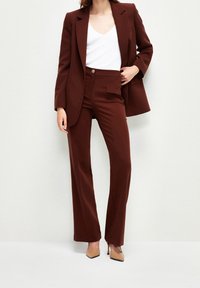 Žena v hnědém ušitém blejzru a odpovídajících kalhotách, s bílým topem ve tvaru V a béžovými špičatými podpatky, stojící na světlém pozadí.