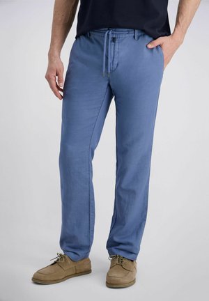 Mann trägt blaue Jogginghose mit Kordelzug und beige Schnürschuhe, steht vor einfachem Hintergrund.