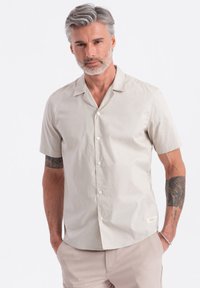 Kurzarm-Hemd aus leichtem Baumwollstoff in Beige, mit einem Reverskragen, normaler Passform und einem kleinen Etikett nahe dem Saum.