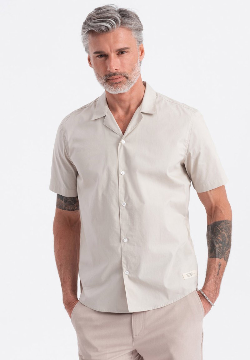 Camisa de botones de manga corta en algodón beige claro, con cuello con muesca, ajuste regular y un pequeño detalle de etiqueta cerca del dobladillo.