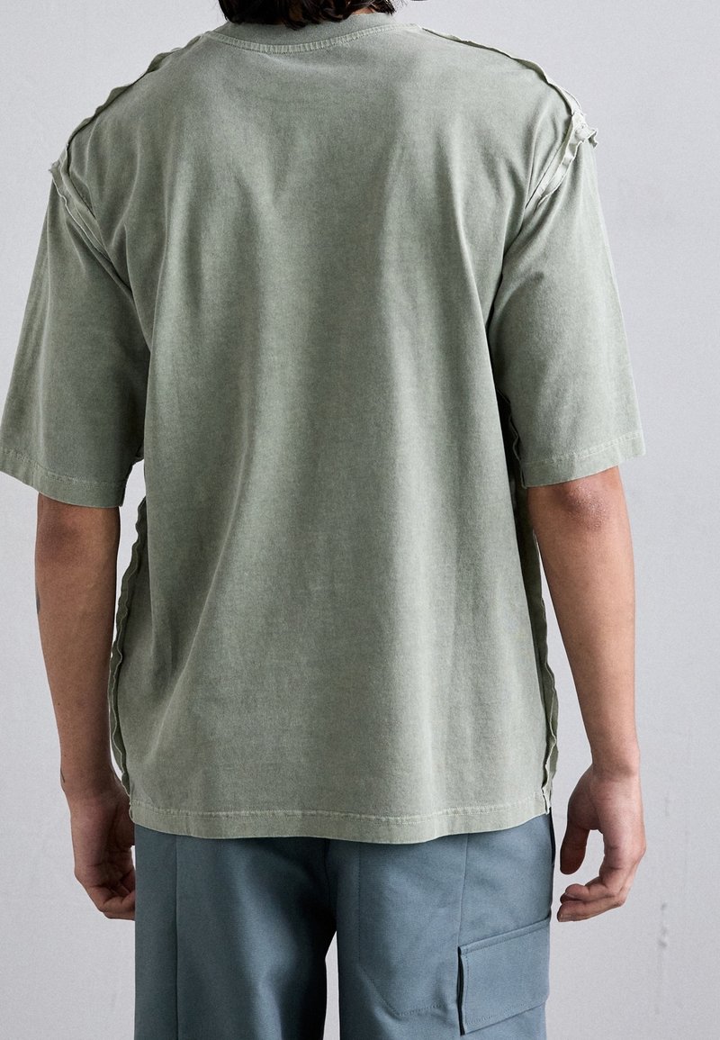 Personne vue de dos portant une chemise verte délavée à manches courtes avec des bords francs aux épaules et un pantalon cargo bleu.
