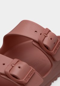 Κοντινό πλάνο ενός ματ σαγιονάρα σε σκόνη ροζ χρώμα με δύο ρυθμιζόμενους ιμάντες με αγκράφες και υφασμένο πέλμα, τονίζοντας την αγκράφα με το ανάγλυφο λογότυπο της μάρκας.