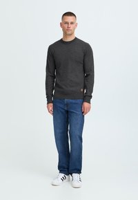 Dunkelgrauer Pullover mit rundem Ausschnitt, enganliegenden Bündchen und einem kleinen Logo-Patch; kombiniert mit blauen Jeans und weißen Sneakern mit schwarzen Streifen.