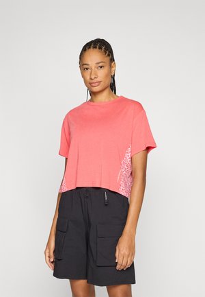 Frau mit geflochtenen Haaren, die ein korallenfarbenes, bauchfreies T-Shirt mit weißen Seitenmustern und schwarze Cargo-Shorts vor einem schlichten Hintergrund trägt.