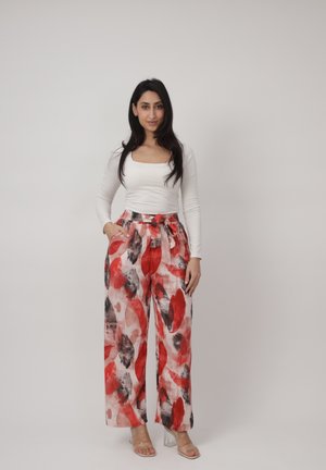 Donna in piedi contro sfondo semplice che indossa una maglia a maniche lunghe bianca e pantaloni a gamba larga e vita alta con stampa astratta rossa e nera.