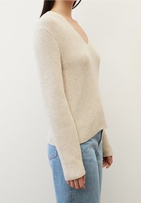 Beżowy sweter dzianinowy z V-neckiem, prążkowaną strukturą i długimi rękawami, w połączeniu z jasnoniebieskimi dżinsami.