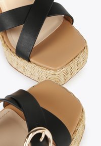 Sandalias de plataforma con tiras cruzadas negras, con una plantilla sintética de color marrón y suela de rafia trenzada. Presenta un diseño de punta cuadrada y un acento metálico.