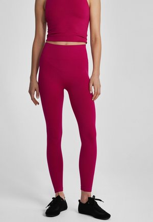 Osoba w ciemnoróżowych legginsach z wysokim stanem i dopasowanym krótkim topie, stojąca w czarnych sportowych butach na jasnoszarym tle.