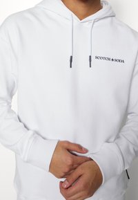 Bílá mikina s kapucí a žebrovanými manžetami. Na hrudi je námořnicky modré logo "SCOTCH & SODA". Měkká tkanina, klasický střih, nastavitelná stahovací šňůrka.