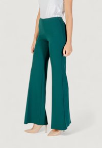Pantaloni a gamba larga color teal realizzati in tessuto lucido, con vita alta e cerniera laterale. Abbinati a tacchi beige per un contrasto.