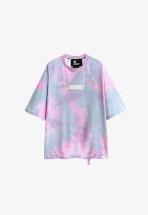 Joven de espaldas con una camisa tie-dye en tonos pastel con alas de lentejuelas de colores, pantalones cortos rojos, calcetines blancos y zapatos beige texturizados.