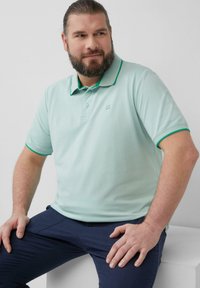 Lichtgroen poloshirt met een plat kraag, in contrast met donkergroene afwerking. Korte mouwen en subtiele geborduurde logo. Draagt met donkerblauwe broeken.