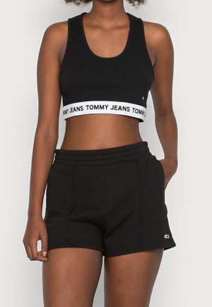 Kobieta nosząca czarny sportowy krótki top z białym paskiem "Tommy Jeans" oraz czarne szorty z małym logo na lewej nodze, pozuje z jedną ręką w kieszeni.