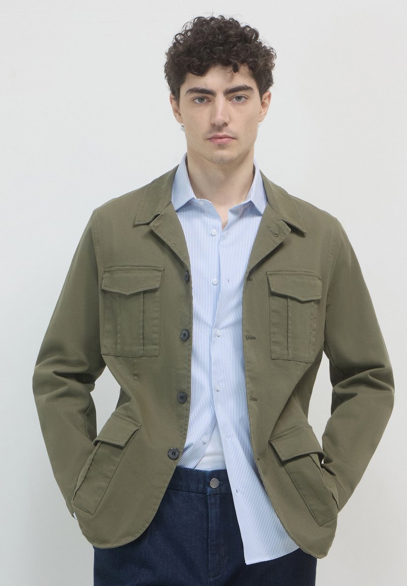 Olivgrüne Jacke mit zwei Brusttaschen, durchgehender Knopfleiste und Kragen, getragen über einem hellblauen gestreiften Hemd.