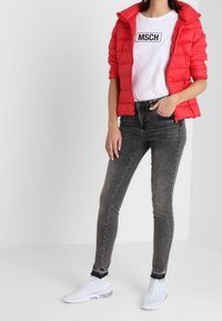 Chaqueta acolchada roja, camiseta blanca con el logo "MSCH", jeans ajustados oscuros y zapatillas blancas con detalle texturizado.