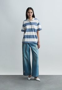 Jeune femme debout devant un fond uni, portant un polo rayé bleu et blanc, un jean large brodé et des talons blancs pointus.