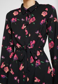 Robe noire à fleurs avec des fleurs roses et violettes vibrantes, manches longues, devant boutonné et détail de taille noué. Texture de tissu douce.