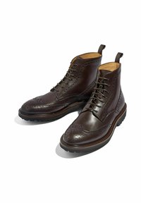 Botas brogue de cuero marrón con perforaciones decorativas, diseño con cordones y tirantes. Cuenta con una suela de goma duradera y costuras en contraste.