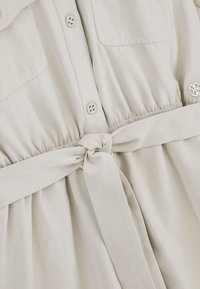 Jasnobeżowa sukienka z przodu na guziki, z dwiema kieszeniami na piersi, elastyczną talią i związanym paskiem. Wykonana z miękkiego, gładkiego materiału.