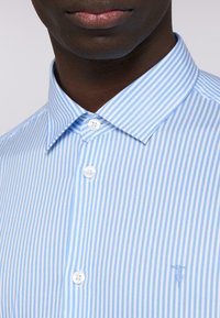 Světle modrá a bílá pruhovaná košile s límcem na knoflíky, která má bílé knoflíky a malé vyšívané logo na hrudi.