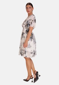 Vestido floral en crema y gris con mangas cortas, cintura ajustada y falda fluida. Acompañado de tacones negros con un acento decorativo.