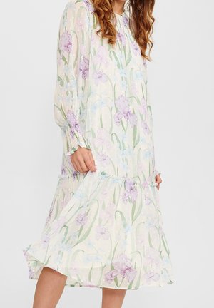 Femme portant une robe midi florale pastel à manches longues avec un ourlet à volants et des poignets smockés sur un fond uni.