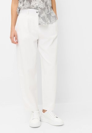 Personne portant un pantalon droit blanc taille haute, des baskets blanches et un haut à motif noir et blanc, une main dans la poche.
