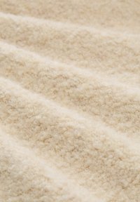 Cremfarbener Stoff mit einer weichen, strukturierten Oberfläche, die aus erhöhten, geschlungenen Fasern besteht, die in sanften, gewellten Linien angeordnet sind.