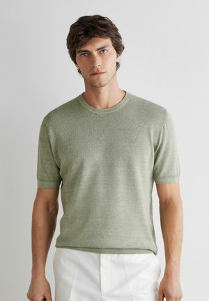 Ung mand med kort brunt hår iført en lysegrøn kortærmet sweater og hvide bukser, stående foran en ensfarvet lys baggrund.