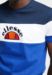 Muž oblečený v tričku s barevným blokováním v modré, bílé a námořnické barvě, s oranžovým a červeným logem ellesse na hrudi.