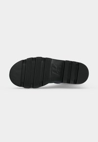 Semelle de chaussure noire avec de larges rainures horizontales et une section centrale texturée sur un fond gris clair.