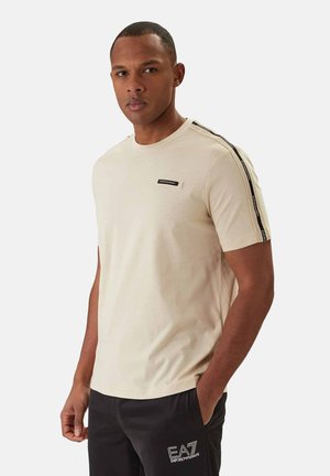 Uomo che indossa una camicia beige a maniche corte con nastro logo a strisce nere sulle maniche e pantaloncini neri con marchio EA7 Emporio Armani.