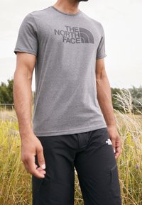 Camiseta gris de manga corta con un escote redondeado y un logo gris oscuro en el pecho. Combinada con pantalones negros, ambientada en un área exterior con césped.