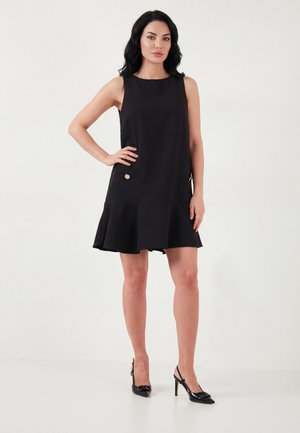 Kvinde står med hånden på hoften iført en ærmeløs sort kjole med guldknapper og sorte højhælede sko på en hvid baggrund.