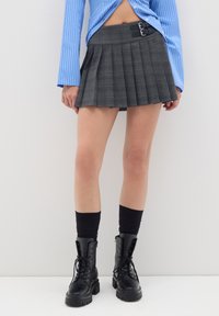 Gonna grigia a pieghe con fibbie in vita, abbinata a stivali militari neri e calze nere sopra il ginocchio. Camicia a righe blu visibile sopra.