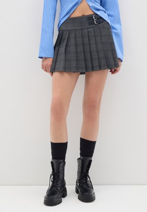 Gonna grigia a pieghe con fibbie in vita, abbinata a stivali militari neri e calze nere sopra il ginocchio. Camicia a righe blu visibile sopra.