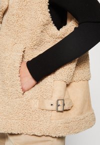 Hand in der Tasche einer beigefarbenen strukturierten Lammfelljacke mit verstellbarem Seitenverschluss über einem schwarzen Rippärmel und beigen Hosen.