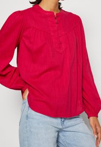 Rosa långärmad blus med hög krage, knappslå, rynkdetaljer och mjuk textur; passar bra med ljusblå denim.